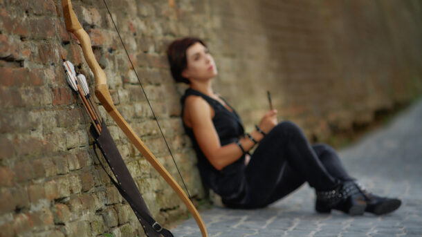 A young brunette woman in black sitting on road, leaning on brick wall, near a resting bow and quiver of arrows